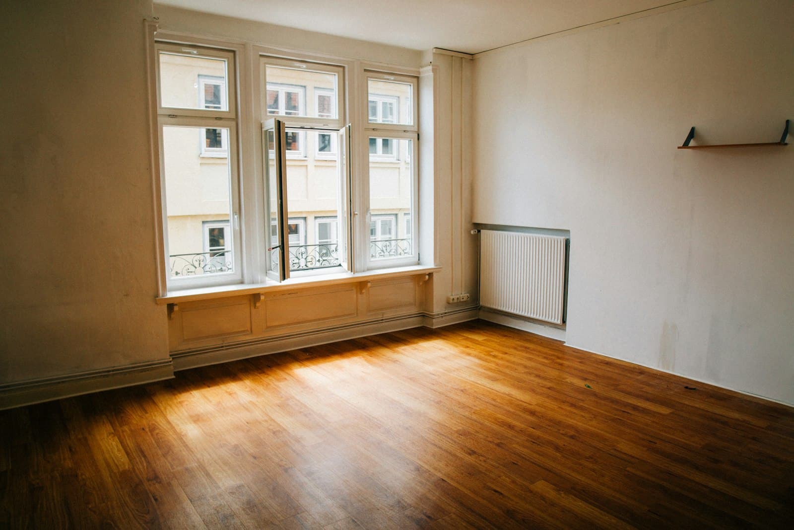 An empty apartment room with afternoon sunlight across the bare wood floor and an open window.